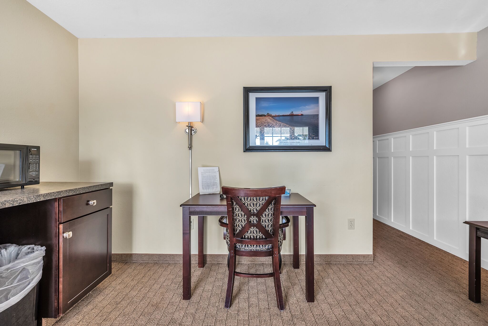 A hotel room with a wooden desk, a chair, a framed picture on the wall, a lamp, and a microwave on the counter.