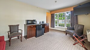 Habitación de hotel con kitchenette, dos sillones estampados, un sofá rojo y un ventanal con cortinas.