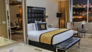 A hotel room with a large bed, a sitting area with a chair and a small table, a large window with a city view, and a bathroom with a bathtub and mirror.
