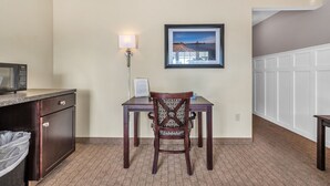 A hotel room with a kitchenette, a dining table with a chair, a framed picture on the wall, and a carpeted floor.