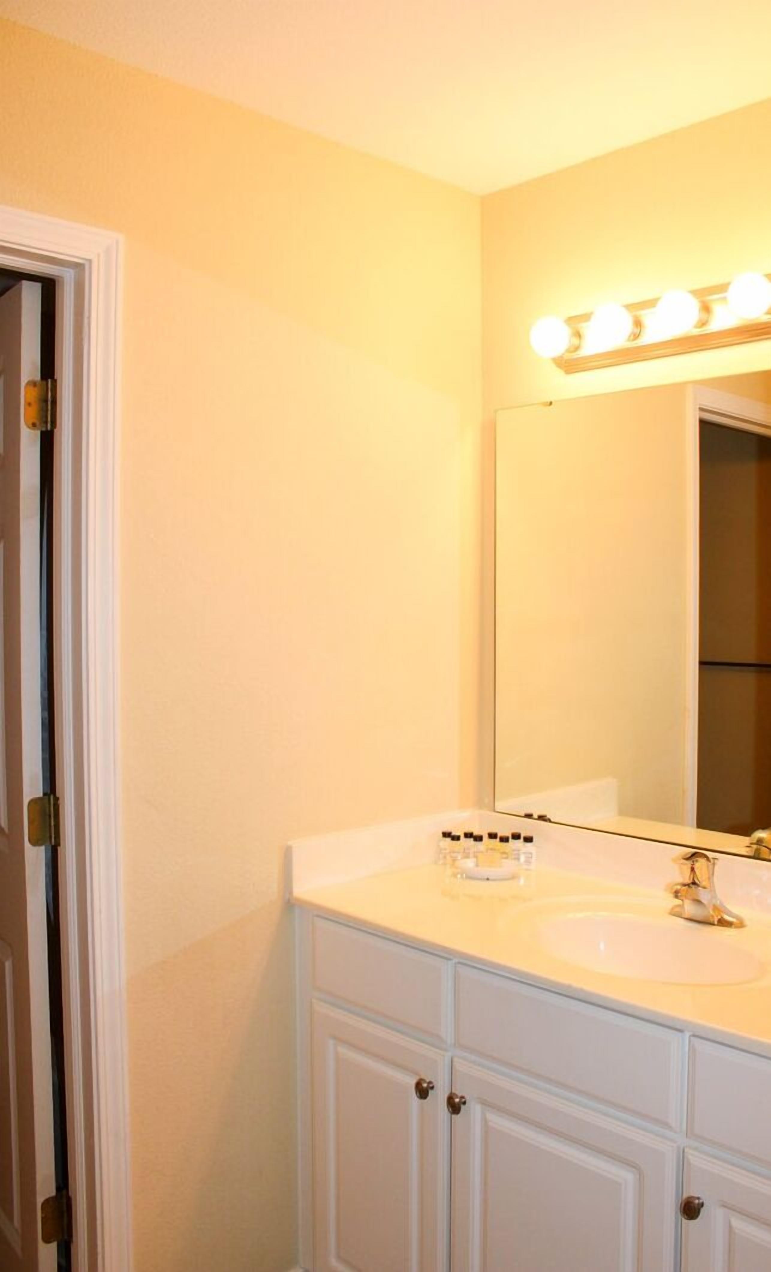 A bathroom with a white vanity, a large mirror with lights, and a door.