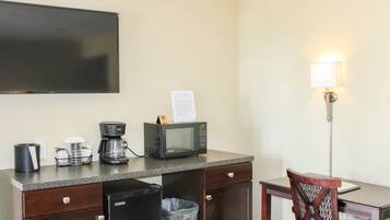 A hotel room with a flat-screen TV, a microwave, a coffee maker, a trash bin, a desk with a chair, and a wall-mounted lamp.