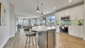 A modern kitchen with white cabinets, a large island with stools, stainless steel appliances, and a green backsplash.