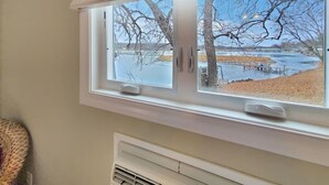 A window with a view of a lake and trees, a white radiator below, and a woven basket to the left. - The Lakeside Lookout on East Lake`s Gingles Point (Arnolds Park)