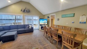 A living room with a sectional sofa, wooden dining table, and chairs, a television, and a view of the outdoors. - The Lakeside Lookout on East Lake`s Gingles Point (Arnolds Park)