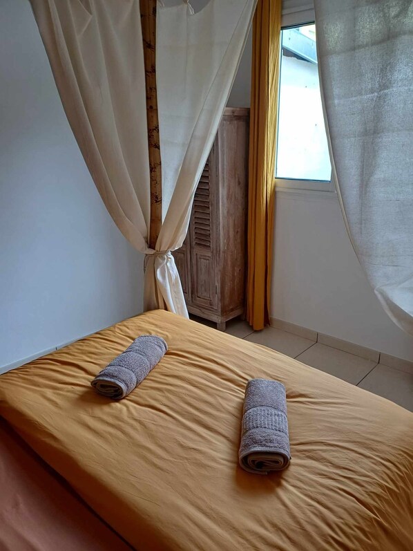 A single bed with a yellow mattress, two rolled-up towels, and a wooden wardrobe in a room with a window and sheer curtains.