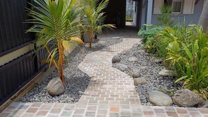 A paved pathway leading to a house with a covered porch, surrounded by a variety of plants and a black fence. - Ô Licuala Villa (Petite Île)