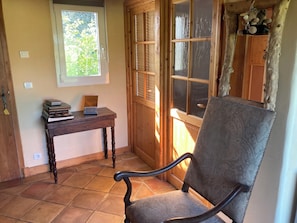 Una habitación con una silla de madera, una mesa con libros y una radio, una ventana con persianas, y una puerta con un llavero colgando.