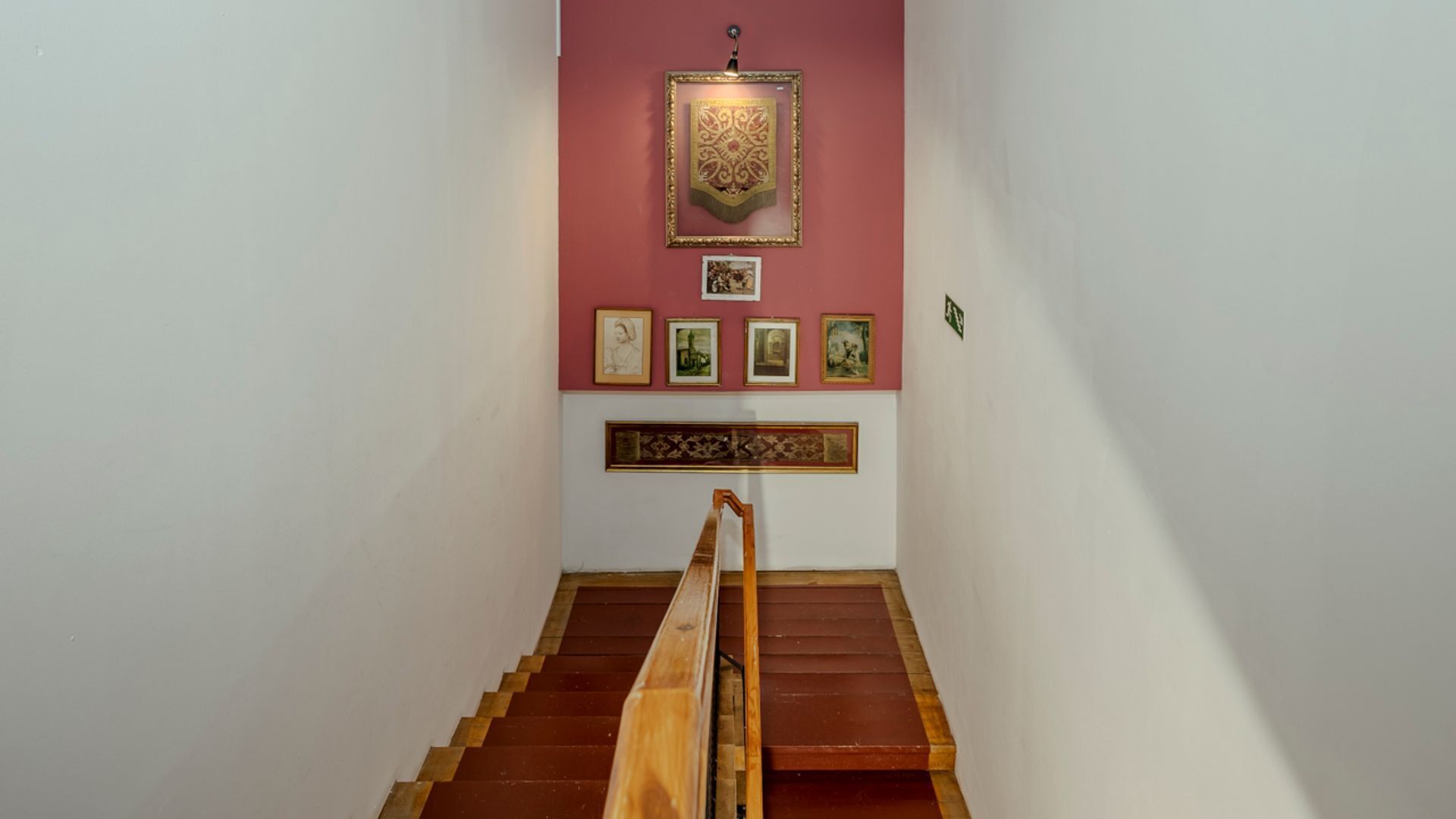 Una escalera de madera con barandilla que lleva a una habitación con pared roja y obras de arte enmarcadas.