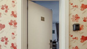 Un couloir avec une porte marquée du numéro « 5 », qui donne sur une pièce contenant un bureau et une chaise, un tableau accroché au mur et un interphone.