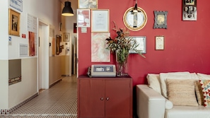 Un couloir avec un sol à motifs, un mur rouge orné de tableaux encadrés et une radio ancienne, ainsi qu’un canapé blanc avec des coussins à motifs.