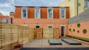 Un edificio de dos pisos con cerca de madera, un pequeño jardín con macetas circulares verdes y una terraza de madera con un banco.