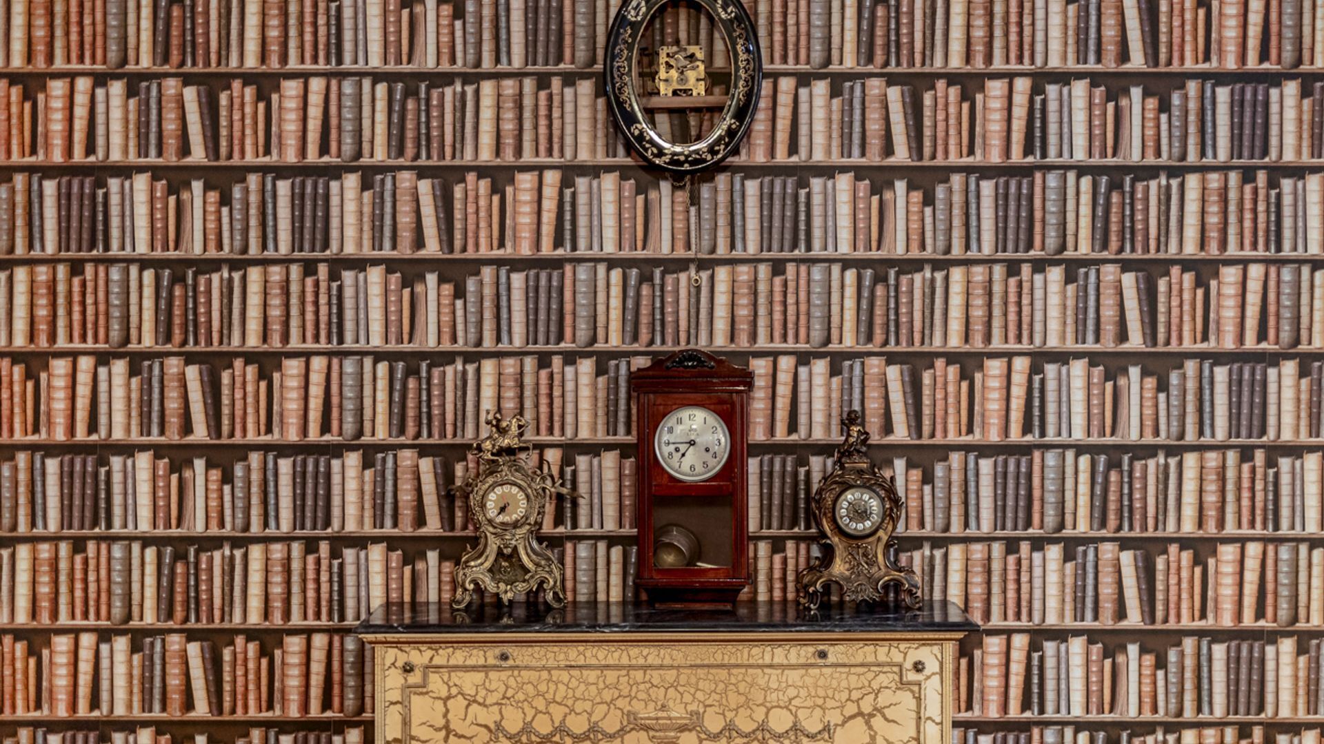 Een bibliotheek met boekenkasten vol boeken, een houten bureau met twee versierde klokken en een lijstspiegel aan de muur.
