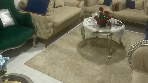 A living room with a flat-screen TV mounted on the wall, two beige sofas with blue cushions, a green armchair, and a coffee table with a vase of flowers. - بيت عطلات (Awlad Saqr)