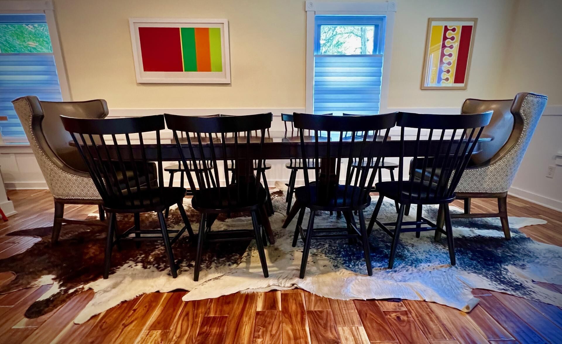 A dining area with a wooden table and eight chairs, a cowhide rug, and two windows with blinds.