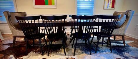 A dining area with a wooden table and eight chairs, a cowhide rug, and two windows with blinds.