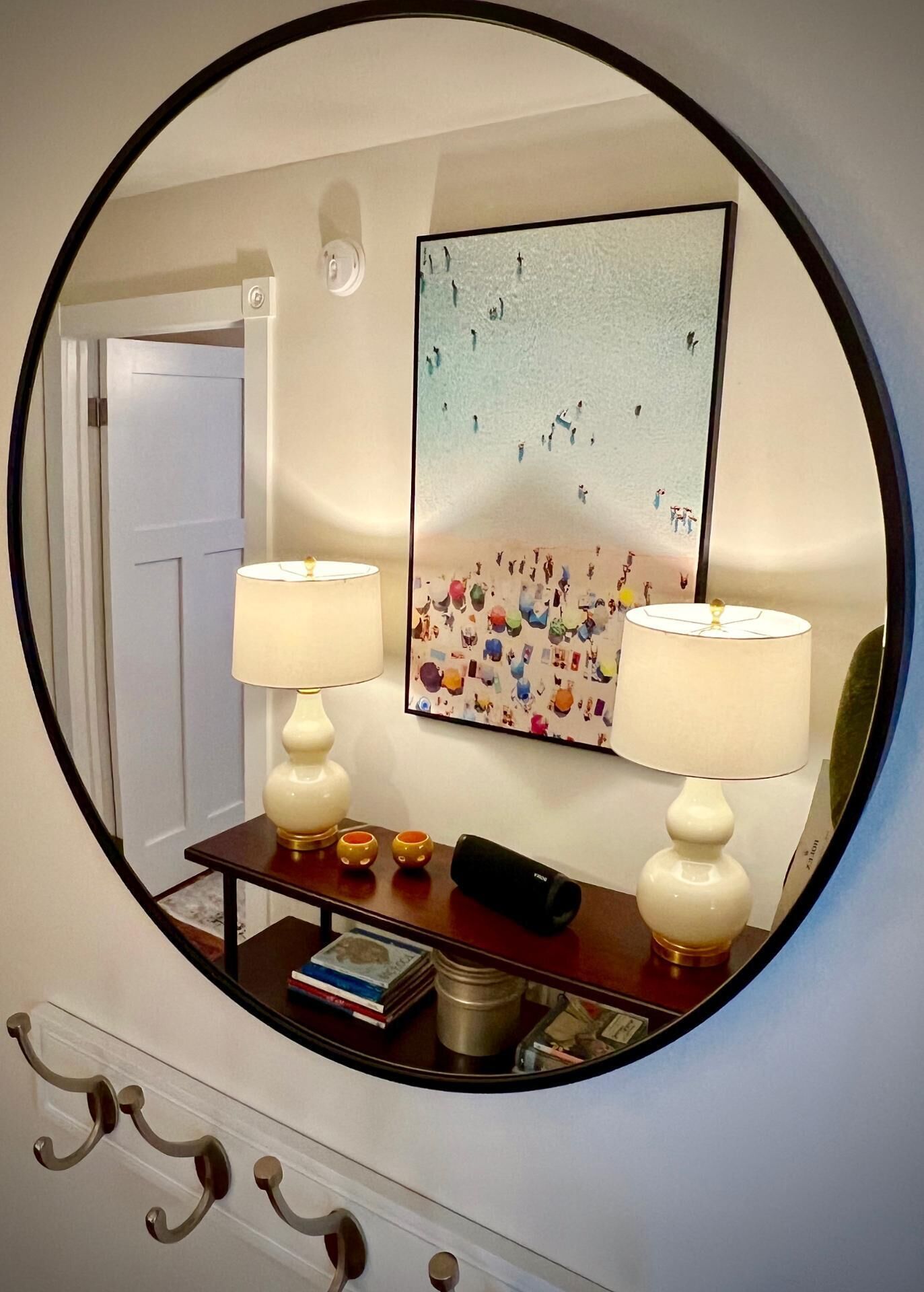 A mirror reflecting a room with two lamps, a framed artwork, and a small table with books and a mug.