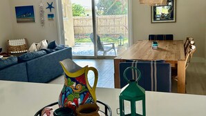 Una sala de estar con un sofá, una mesa de comedor con sillas, una mesa de centro con adornos y un ventanal corredizo que da a un jardín trasero.