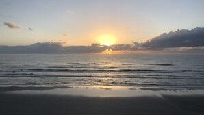 Escena de playa con el sol poniéndose sobre el océano, olas tranquilas y cielo despejado.