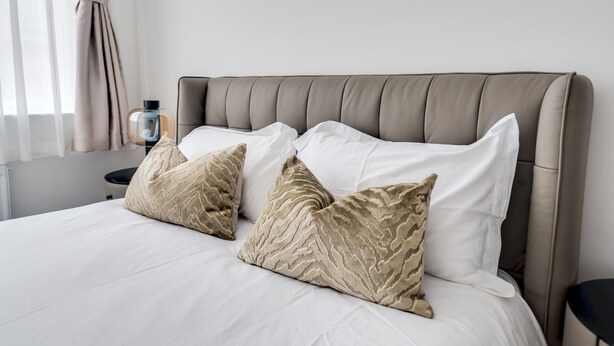 A bed with white linens and two beige patterned pillows, a bedside table with a bottle, and a window with curtains. - Tranquil 4-Bed Oasis Modern & Chic (Oxford)