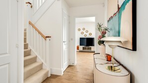 A hallway with a staircase, a TV, a vase with flowers, and a lamp on a console table. - Tranquil 4-Bed Oasis Modern & Chic (Oxford)