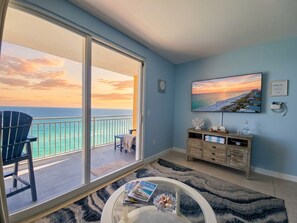Ein Zimmer mit einer großen Schiebetür, die auf einen Balkon mit Meerblick führt. Es gibt einen runden Tisch mit einer Zeitschrift und Dekorationselementen, eine Holzkommode und ein gerahmtes Bild an der Wand.