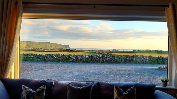 A living room with a large window offering a view of a rural landscape, including fields, a stone wall, and distant hills. - Cozy semi detached cottage 5 mins drive from Doolin. Cliffs of Moher views! (Doolin)