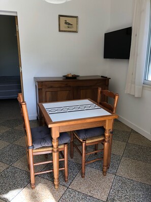 A dining area with a wooden table and chairs, a small black bowl with fruits on a wooden cabinet, a framed picture on the wall, and a flat-screen TV mounted above the cabinet. - Petite Maison en Provence (Arles)