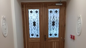 Un couloir avec deux portes en bois ornées de panneaux de verre décoratifs, un mur blanc sur lequel est fixée une plaque et un sol carrelé.