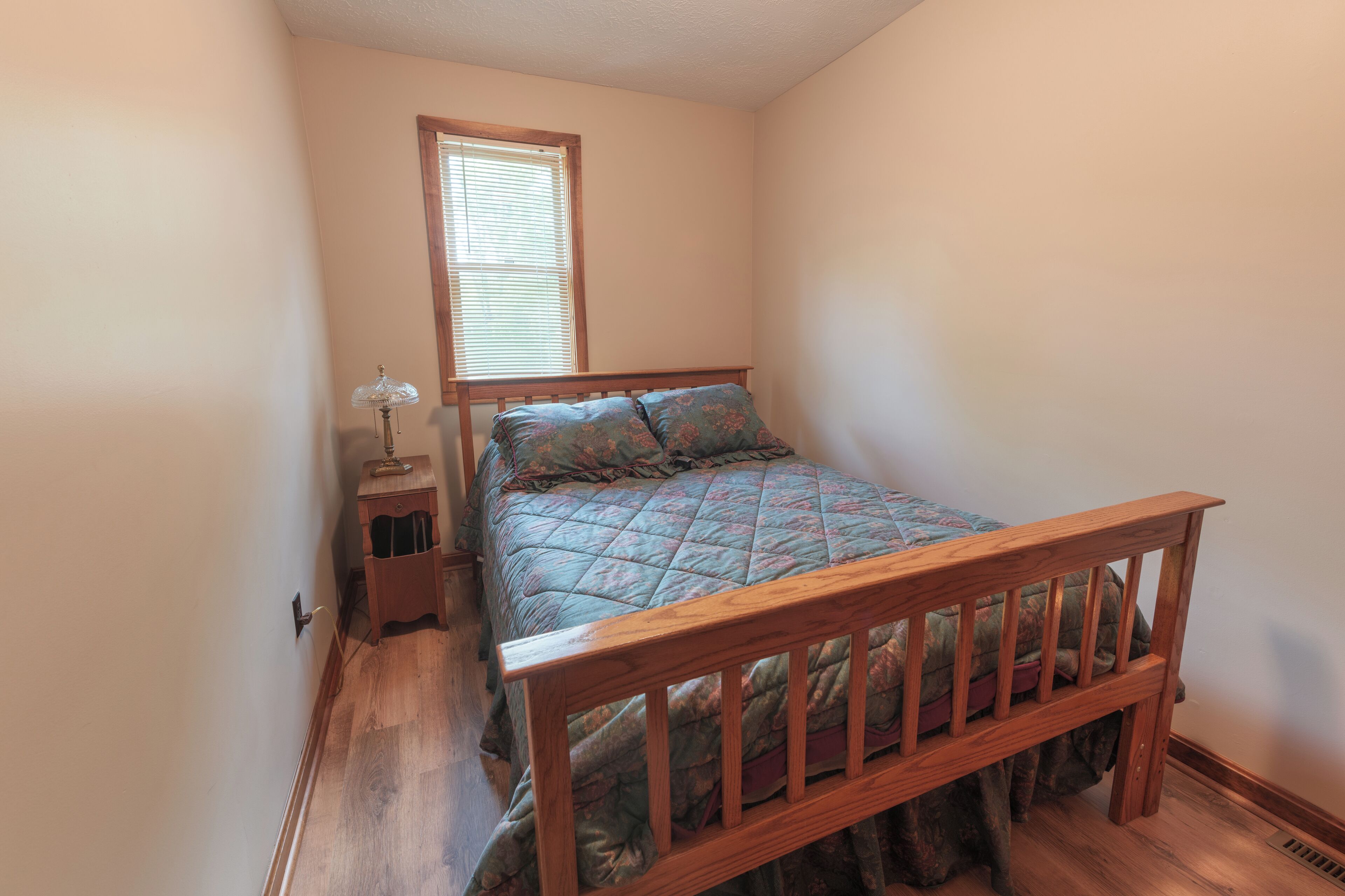 A bedroom with a wooden bed frame, a nightstand with a lamp, and a window with blinds.