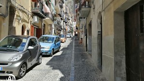 Una estrecha calle empedrada flanqueada por edificios de varios pisos, coches aparcados y balcones con plantas y ropa tendida.