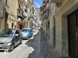 Una estrecha calle empedrada flanqueada por edificios de varios pisos, coches aparcados y balcones con plantas y ropa tendida.