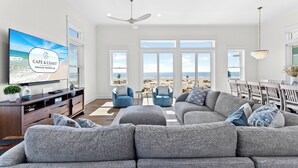 Un salon spacieux avec un grand canapé d’angle gris, deux fauteuils bleus, une table basse en bois et un téléviseur à écran plat fixé au mur.