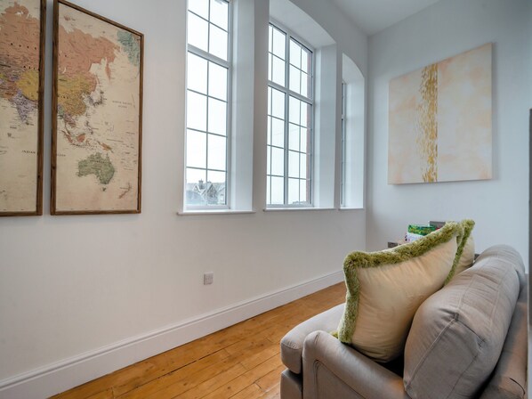 Una habitación con piso de madera, un sofá gris con un cojín verde y blanco, y dos ventanas amplias.