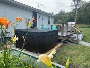 A hot tub in front of a house with a deck, surrounded by greenery and flowers.