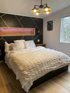 A modern bedroom with a large bed, a headboard with built-in lighting, a bedside table, and a window with a view of trees.