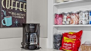 Et køkkenområde med kaffemaskine, et skilt med kaffemotiv på væggen og hylder fyldt med køkkenudstyr og madvarer.