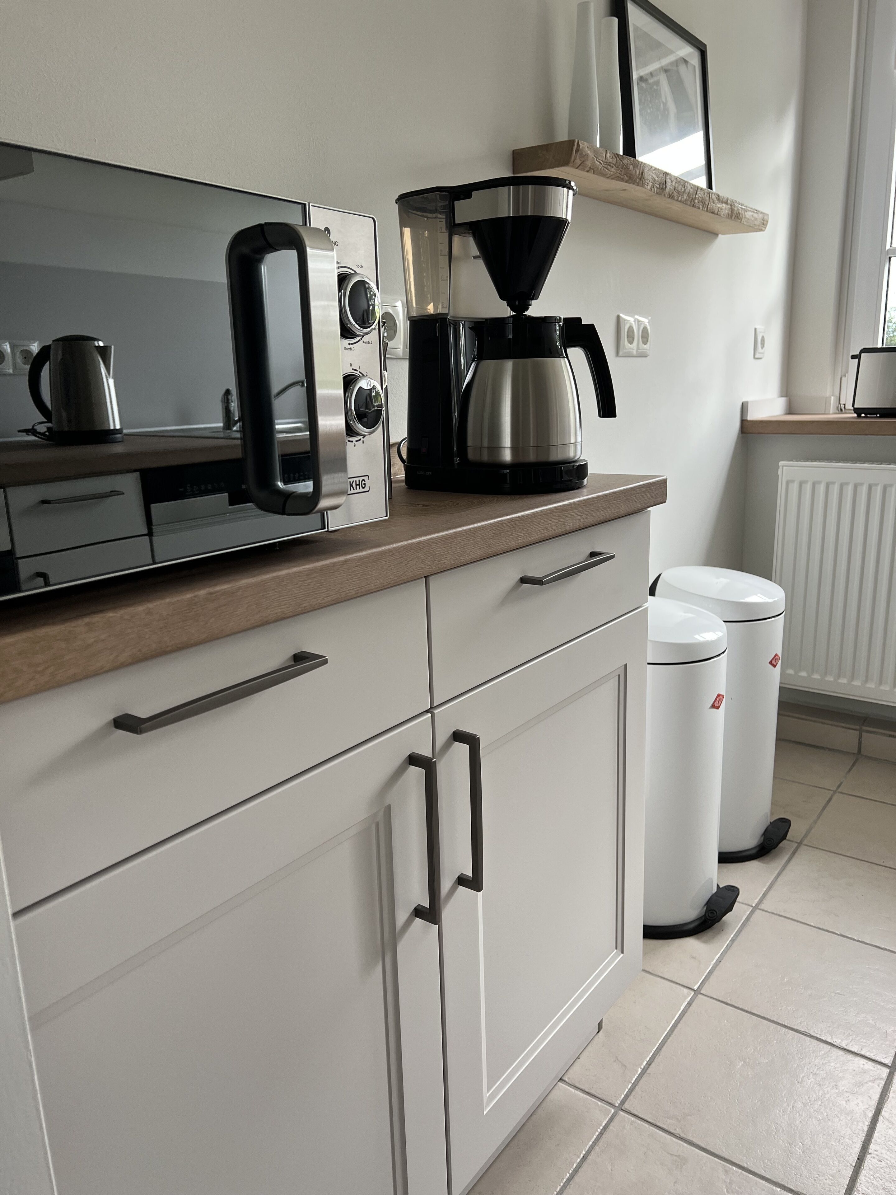 A modern kitchen with a coffee maker, kettle, and a built-in oven. There are white cabinets with black handles and a wooden countertop.