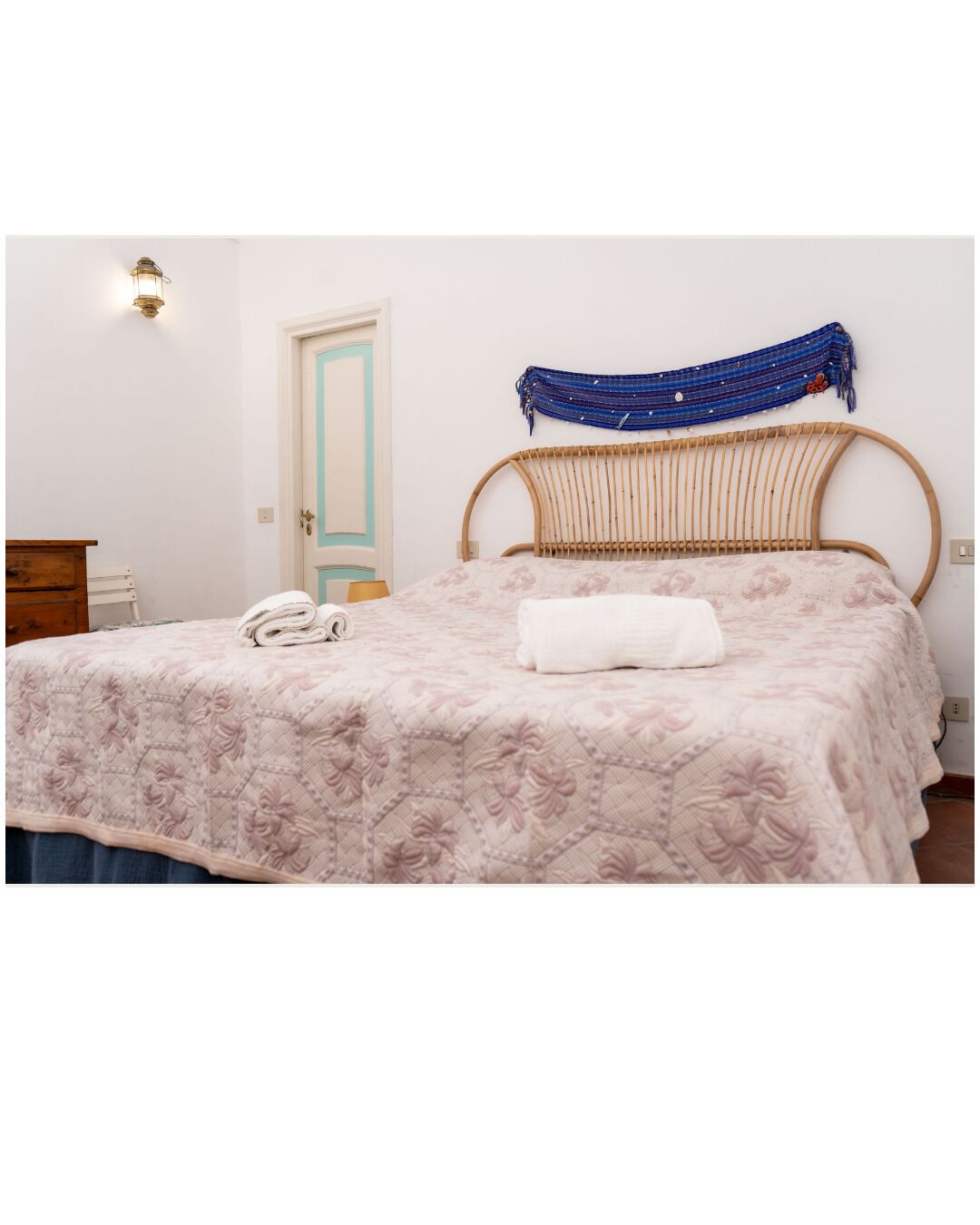 Un dormitorio con una cama, cabecera de madera, lámpara de pared, una puerta y un pequeño aparador de madera.
