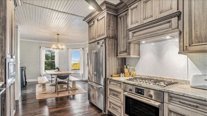 A kitchen with wooden cabinets, stainless steel appliances, a dining area with a table and chairs, and a view through a window. - Lake Michigan Sunset Views from updated Macatawa home w/private beach access  (Holland)