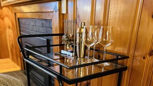 Ein schwarzer Metallwagen mit mehreren Weingläsern und einer Flasche, platziert auf einem Holzboden nahe eines Kamins.