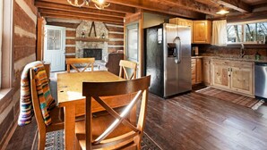 A rustic cabin interior with a dining area, kitchen, and stone fireplace.