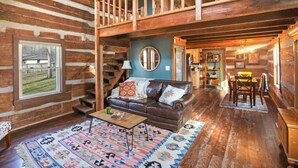 A log cabin interior with a leather sofa, a dining table, and a staircase leading to an upper level.