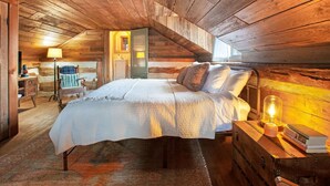 A cozy attic bedroom with a wooden ceiling, a bed with a white bedspread, and a nightstand with a lamp.