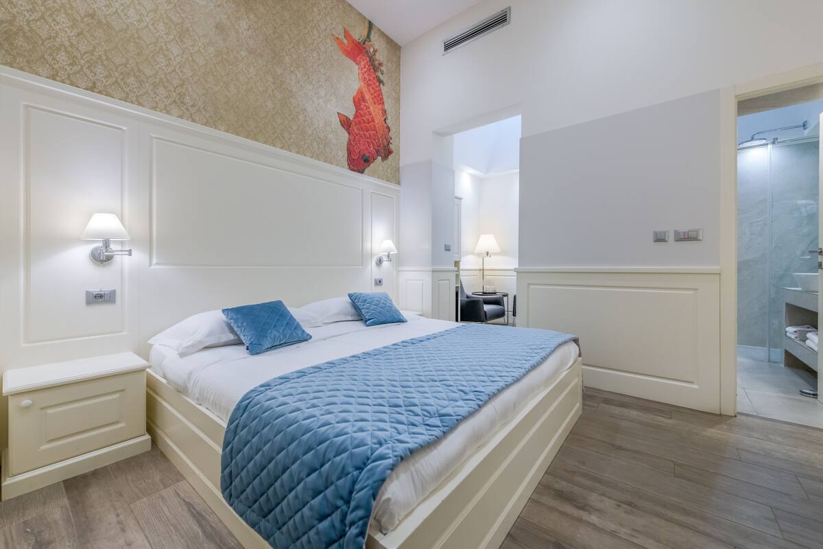 Un dormitorio con cama, lámparas de noche, mesita de noche, espejo y baño visible a través de una puerta abierta.