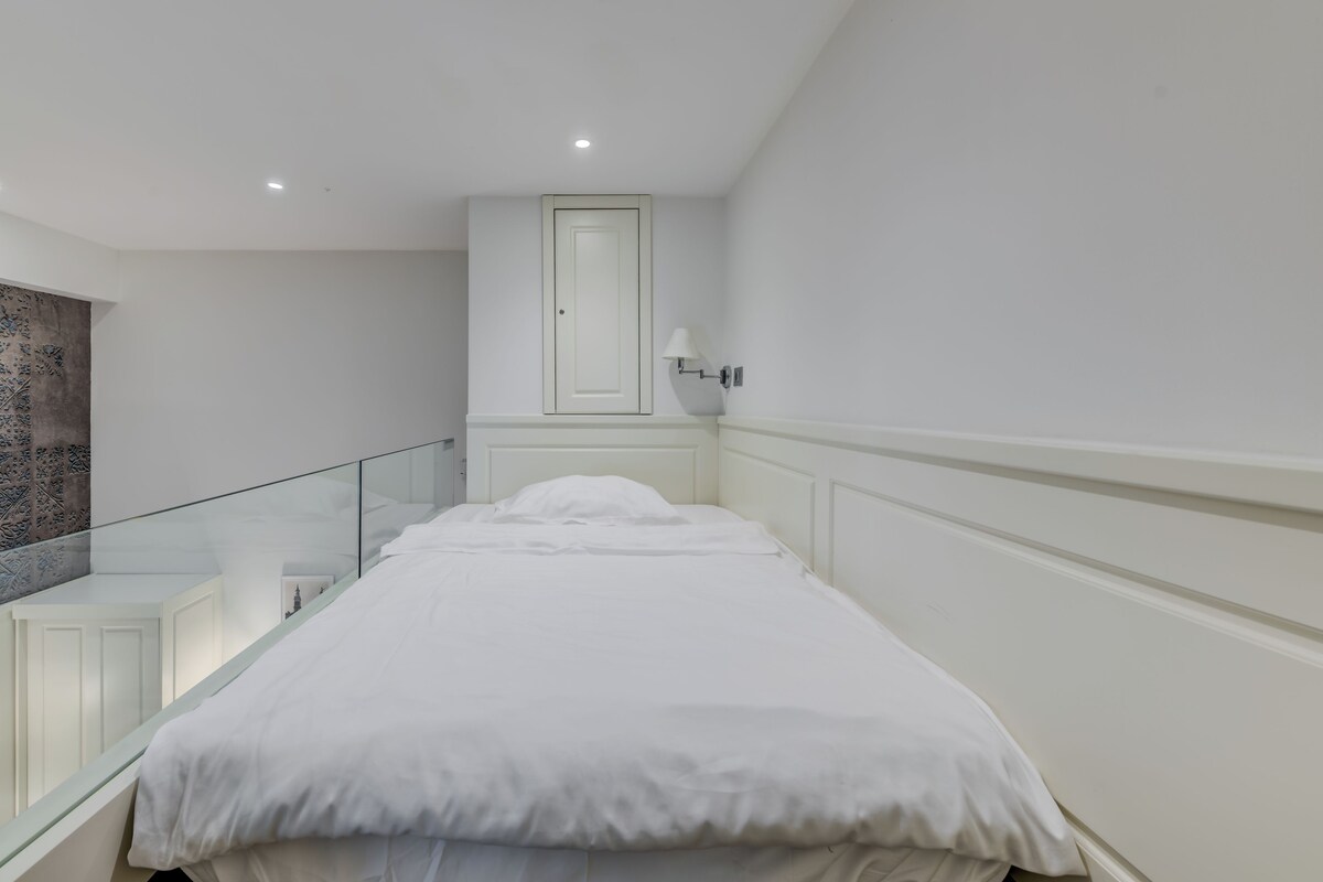 Un dormitorio moderno con una cama individual, barandilla de cristal, una puerta blanca y una lámpara fijada a la pared.