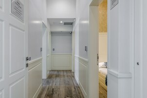 A narrow hallway with white walls and wooden flooring, leading to a room with a bed.