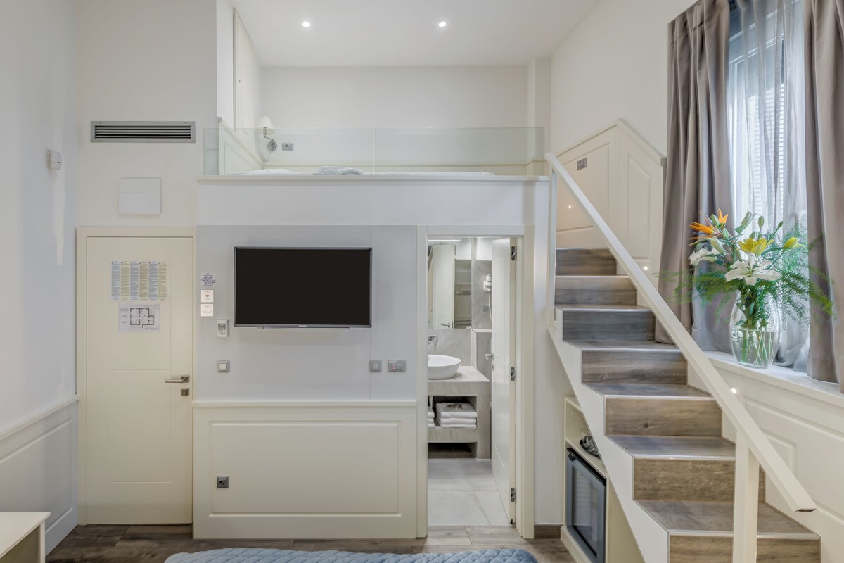 Habitación de hotel moderna con televisión de pantalla plana montada en la pared, una escalera que conduce a un nivel superior y vistas al baño.