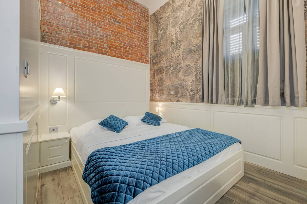 Un dormitorio con pared de ladrillo, un ventanal con cortinas, una cama blanca con ropa de cama azul y una mesita de noche con lámpara.