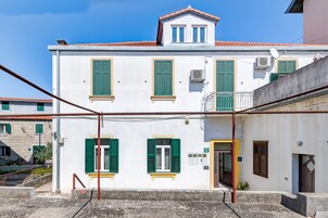 A two-story building with green shutters, a balcony, and a small staircase leading to the entrance.